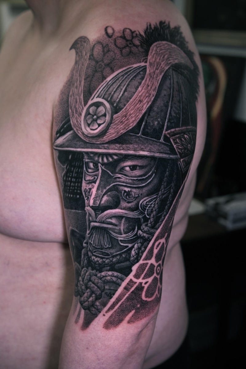 Black & Grey Tattoo am Oberarm: Samurai mit Kabuto-Helm und Gesichtsmaske, Kirschblüten und geflochtene Seildetails