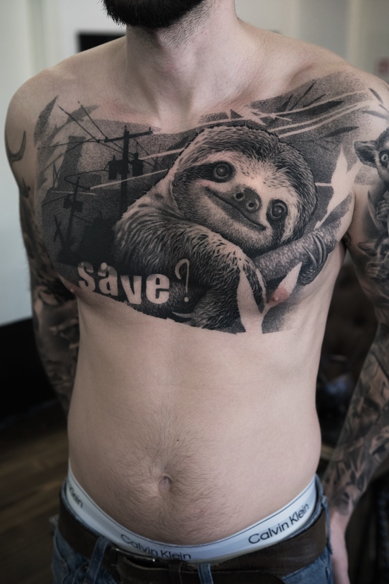 Brust-Tattoo in Black & Grey: Faultier an einem Ast vor grafischem Hintergrund mit Strommasten und dem Schriftzug „Save?“